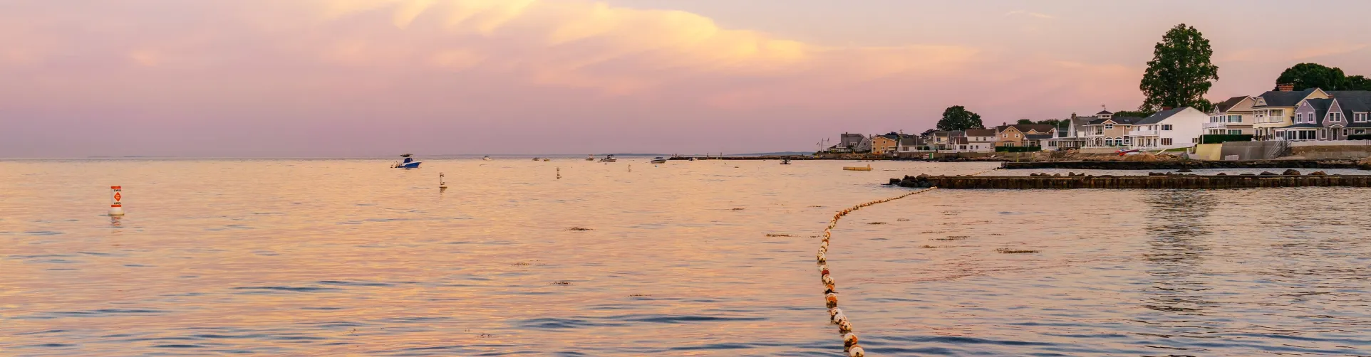 A picturesque coastal scene at sunset with calm water, a long line of buoys, and houses along the shore