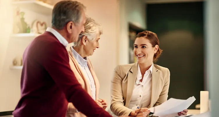 A smiling female financial advisor seated at a counter, reviewing documents and talking with an older couple who are conversing with her