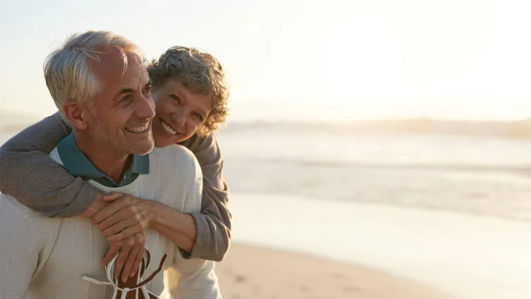 An older man giving an older woman a piggyback ride on a beach, both smiling and looking happy